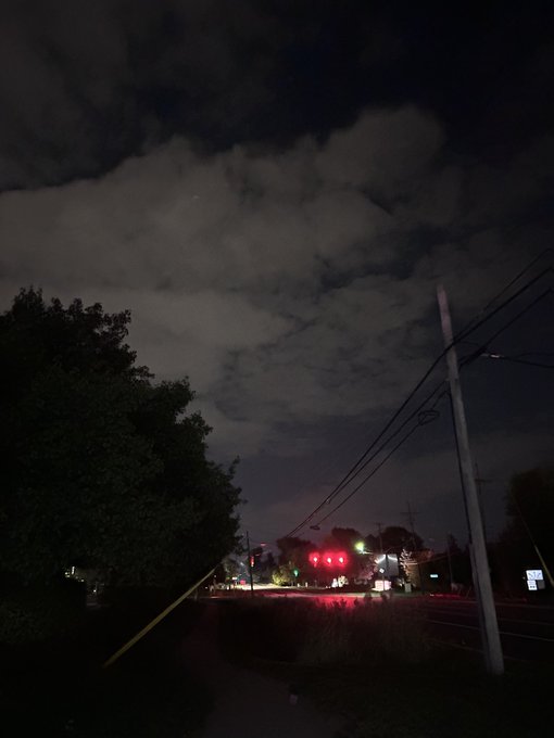 A nighttime photo showing a dark, cloudy sky above a street with a traffic light glowing red, utility poles with power lines, and trees silhouetted in the foreground.