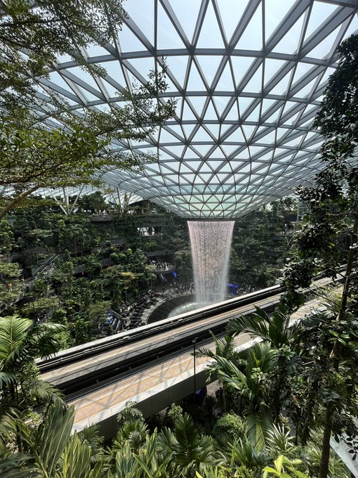 An indoor view of a large indoor waterfall at Jewel Changi Airport, surrounded by lush greenery under a geometric glass dome ceiling.
