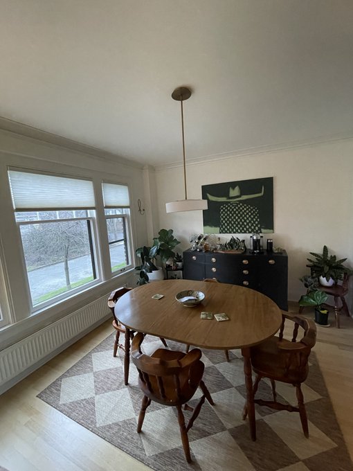 An interior view of a dining room with a wooden oval table, four chairs on a geometric rug, a dark dresser, plants, and large windows.