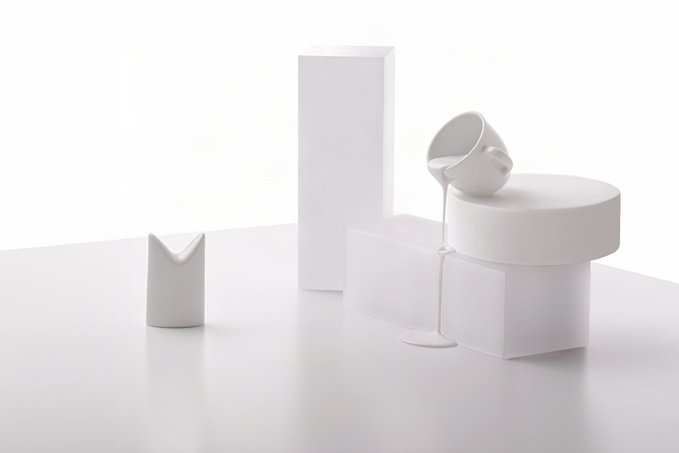 A minimalist 3D render showing white geometric shapes—cubes, cylinders, and a tilted cup—with white liquid pouring out onto a white surface.