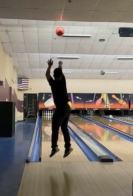 A person mid-jump at a bowling alley, arms raised to release a red bowling ball down the lane, with an American flag and pins visible in the background.