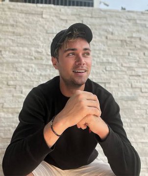 A portrait of a young man wearing a black cap and black sweatshirt, smiling while resting his chin on his hands in front of a white brick wall.