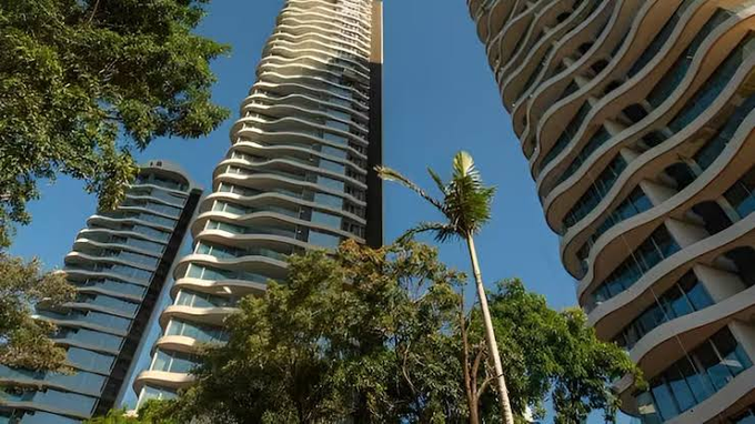 A low-angle photograph of modern high-rise apartment buildings with curved, wavy balconies, framed by palm trees and lush greenery against a clear blue sky.
