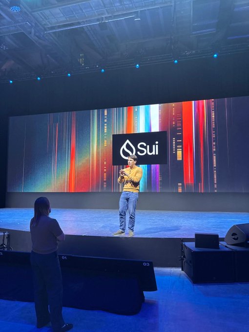 A presenter stands on a stage at a Sui conference, with the 'Sui' logo displayed on a large screen behind them.