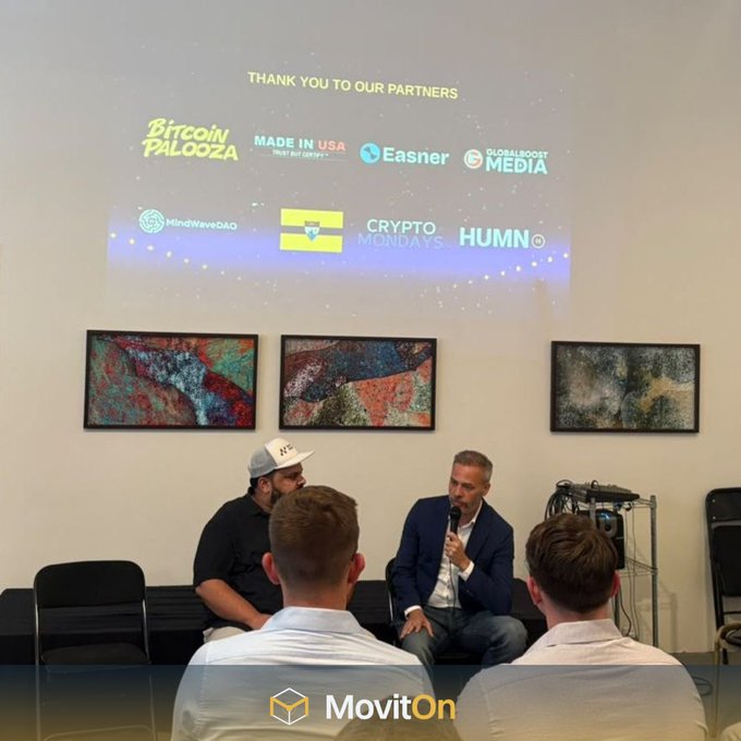 Two men sit on a stage during a panel discussion, with a projected slide thanking partners including 'Bitcoin Palooza', 'Easner', 'MindWaveDAO', 'Crypto Mondays', and 'HUMN', above framed artwork.