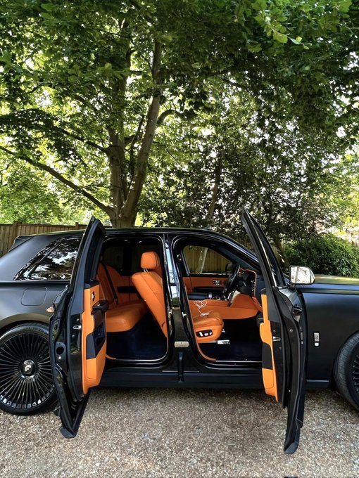 The image features an orange Rolls Royce car parked next to another car with its doors open. The scene takes place outdoors under trees, suggesting that they are at a park or a similar outdoor location. The main focus of the image is on the two cars, and there's no text present in the image.