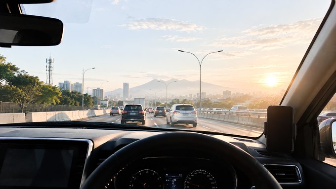 A view from inside a car driving on a highway at sunset, with a steering wheel and dashboard visible in the foreground.