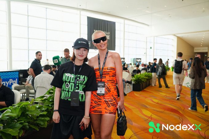 Two women posing together at an indoor conference venue, one wearing a black NodeX t-shirt and the other in an orange NodeX strapless dress, with the NodeX logo and watermark visible in the corner.