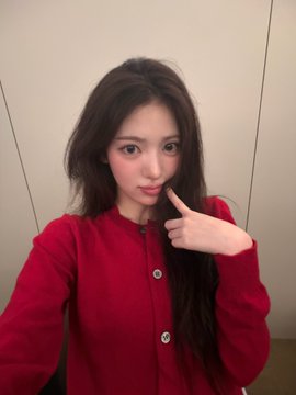 A selfie of a young woman with long dark hair wearing a red cardigan, pointing to her cheek against a plain wall background.