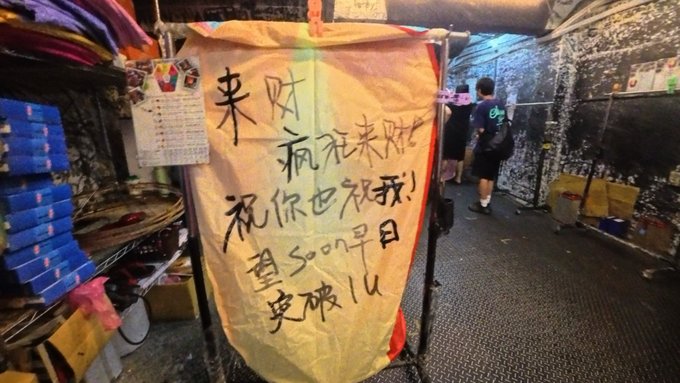 A person stands near a stall with a colorful banner displaying handwritten Chinese characters that translate to "Come get rich, wish you get rich too! I wish for Soon ($SOON) to break 1u soon," expressing the maker's financial goal. The setup, located in what appears to be a market stall, reflects their aspiration for the $SOON token to reach $1, tying personal ambition to the cryptocurrency mentioned in the accompanying post.