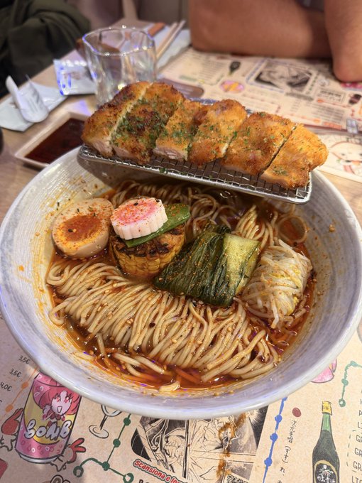 A bowl of spicy ramen noodles topped with green onions, a seasoned egg, and other garnishes, accompanied by a wire rack holding slices of toasted garlic bread on a comic-themed table mat.