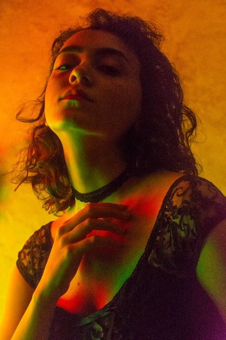 A young woman with her eyes closed and head tilted back poses dramatically under moody green and orange lighting, wearing a black lace top and choker, in a stylized portrait evoking the mystical "witch girl" aesthetic referenced in the accompanying Tezos NFT listing.