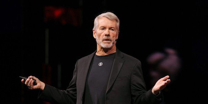 An older man with gray hair and a beard wearing a black blazer and black t-shirt speaking on stage with his arms outstretched.