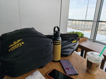 A "Binance" branded bag sits next to a laptop, phone, iced coffee, and water bottle on a table by a window, capturing a travel-ready workspace setup. The Binance bags and travel gadgets reflect a journey to Bali for the Binance Club House event at Coinfest Asia 2026.