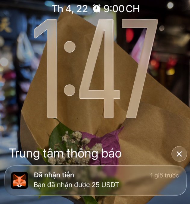 A phone lock screen at 1:47 showing a notification in Vietnamese ('Trung tâm thông báo') confirming receipt of 25 USDT, overlaid on a background of a bouquet wrapped in brown paper.