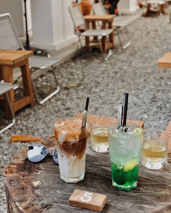 Four drinks on a wooden stump table marked '5' at an outdoor café, including iced coffee and a green cocktail, with chairs in the background.