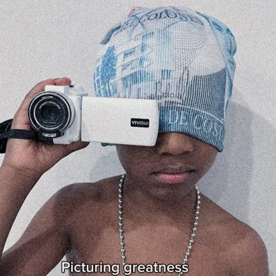 A shirtless person wearing a white and blue cap and a chain necklace, holding a white Vivitar camcorder up to their face, with the text 'Picturing greatness' at the bottom.