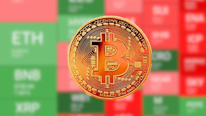 A close-up of a physical gold Bitcoin token positioned in front of a blurred stock market-style price board showing 'ETH' and other cryptocurrency listings on green and red backgrounds.