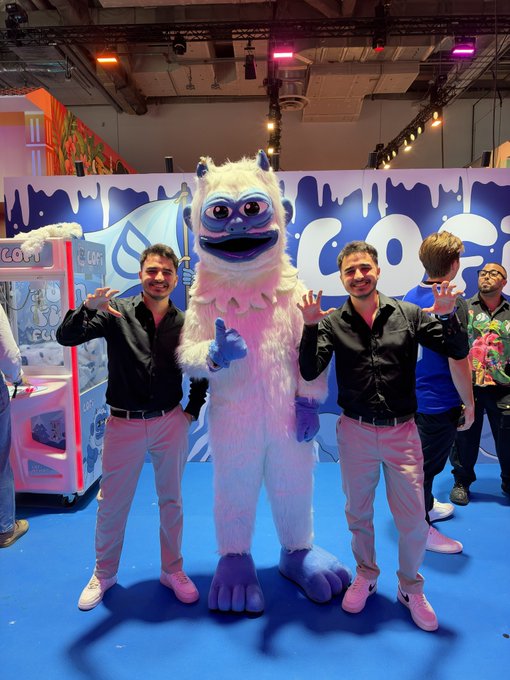 Two men in black shirts and white pants pose on either side of a fluffy white and blue mascot character at what appears to be a convention booth with claw machines.