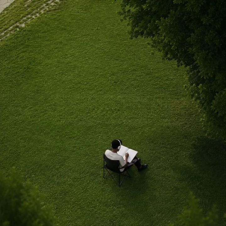 A solitary figure wearing headphones sits in a folding chair on a vast expanse of green grass, sketching in a notebook surrounded by trees. The image evokes the "go offline" theme, illustrating peaceful disconnection while the Crypto.com app keeps watch on the markets in the background.