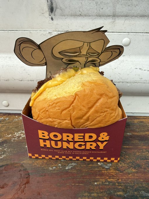 A hamburger in a maroon and yellow checkered box labeled 'BORED & HUNGRY' with the tagline 'BORED APE YACHT CLUB NFT THEMED BURGER RESTAURANT SINCE DSRX IN CALIFORNIA,' featuring a bored ape illustration on the back flap.