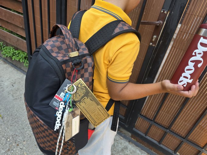 A person wearing a yellow shirt and a checkered backpack holds a maroon water bottle labeled "Decods," with decorative tags and a keychain hanging from the bag.