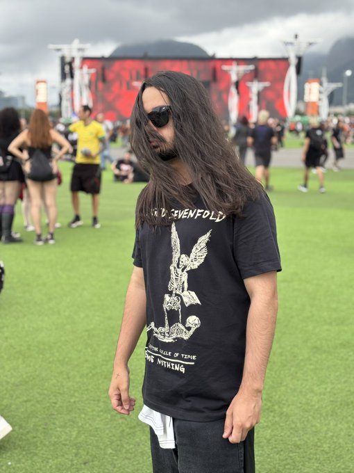 A long-haired man wearing sunglasses and a "Sevenfold" band t-shirt stands on a grassy field at what appears to be the Rock in Rio festival, with a stage and crowd visible in the background. The caption suggests he traveled to Rio specifically to see the band Shadows (likely Avenged Sevenfold), hoping for a memorable encounter.