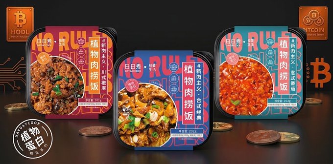 Three self-heating rice bowls branded "HODL Microwave Food" and "BITCOIN Sponsored Food" sit on a wooden surface beside scattered coins, featuring "Plant-Based Meat Rice" flavors including 川式 (Sichuan) and 台式经典 (Taiwanese Classic) in 202g portions, with packaging labeled as "PANDA COOK" — humorously pairing ready-meals with cryptocurrency-themed branding to visually reinforce DayDayCook's expansion into a Bitcoin reserve strategy.