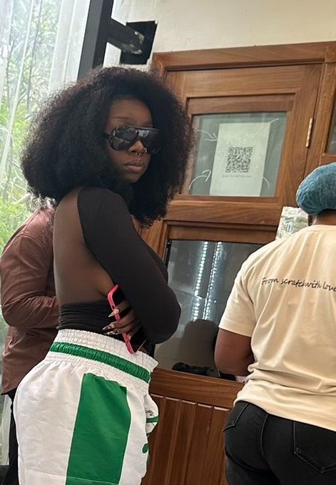 A woman with a voluminous afro wearing sunglasses, a black long-sleeve top, and white pants with a green stripe poses for a photo in what appears to be a café, with a person in a cream 'Fom scratch with love' t-shirt nearby.