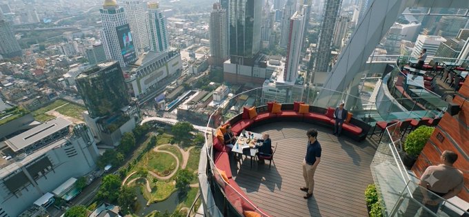 An aerial view from a high-rise rooftop bar/lounge where patrons are seated at tables, overlooking a dense urban cityscape with numerous skyscrapers and a roundabout below.