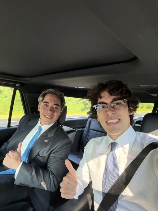 Two men in business suits smiling and giving thumbs up while sitting in the back seat of a moving car.