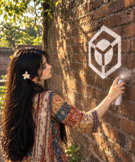 A woman with long dark hair and a flower tucked behind her ear, wearing a floral dress, uses a spray can to paint a white geometric logo on a brick wall.