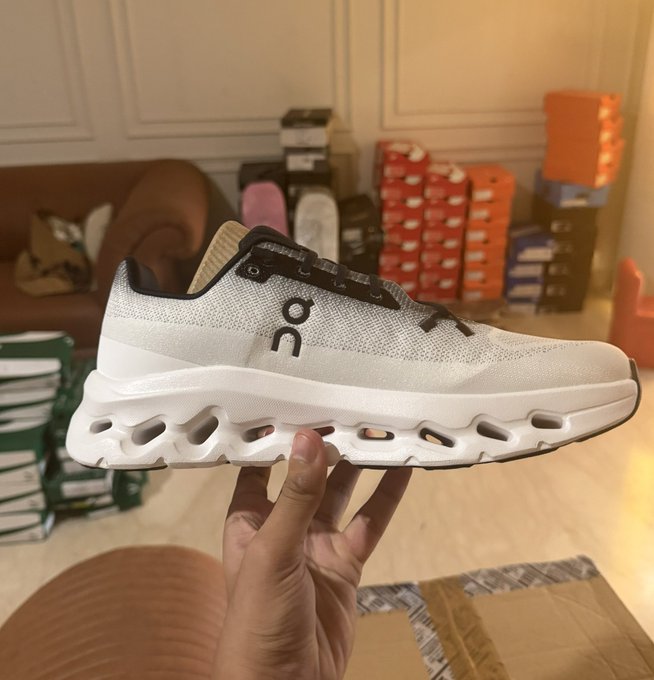 A hand holding up a white On running shoe in front of a background of stacked shoe boxes.
