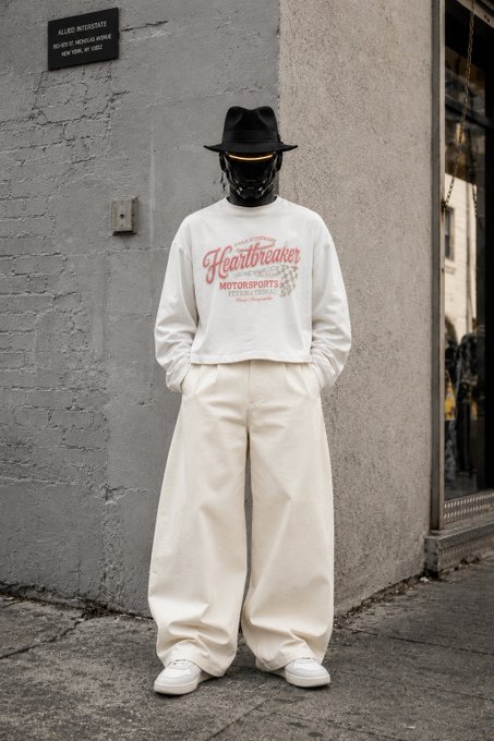 A figure stands on a sidewalk in front of a gray wall, wearing a black fedora, a black face covering that obscures their features, an oversized white sweatshirt with "Heartbreaker Motorsports" printed in red and orange lettering, baggy cream-colored pants, and white sneakers. The streetwear look ties into the post's "GM CT HAPPYTUESDAY ♟️" vibe, with the chess king symbol hinting at the bold, strategic, and statement-making style of the all-white outfit paired with edgy accessories.
