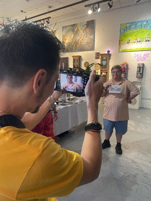 A behind-the-scenes photo of someone filming another person on a smartphone at what appears to be an art gallery or studio space, with colorful paintings on the white brick walls.