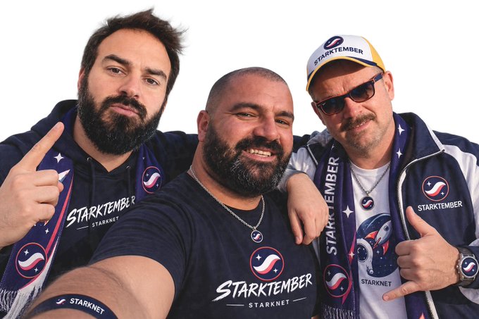 Three smiling men posing together, wearing 'STARKTEMBER - STARKNET' branded apparel, with one taking a selfie showing off a Starknet cap and merchandise.