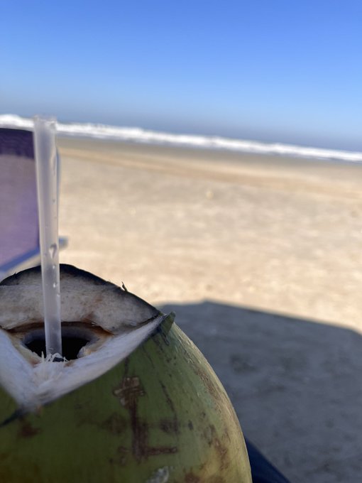 A photo taken at the beach showing a fresh green coconut with a clear plastic straw inserted, with sandy shore and ocean waves visible in the blurred background under a clear blue sky.