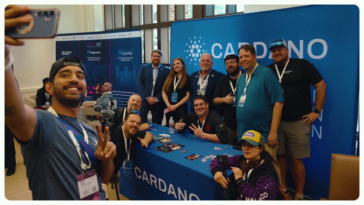The image shows a group of people posing for a photo at an event with a blue backdrop featuring the Cardano logo. The attendees are wearing business attire, suggesting that they might be professionals or industry experts. There are several individuals standing around a table in front of them, and some are holding cell phones to capture the moment. The post about the unified global events marketing strategy highlights how the Cardano brand was brought to the whole world through collaboration with various organizations and the use of funding from Treasury.