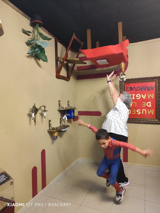 A woman and a child pose playfully in an upside-down museum exhibit, with chairs mounted to the ceiling and a framed "Museo de la Magia Aranjuez" sign displayed on the wall. The image captures the whimsical trick-of-the-eye effect of the room, fitting perfectly with the #aranjuez Museo de la Magia theme.