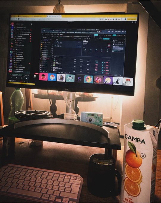 A cozy desk setup featuring a wide LG UltraGear monitor displaying a crypto trading interface, accompanied by a white keyboard, a small camera, and a carton of orange Campa juice.