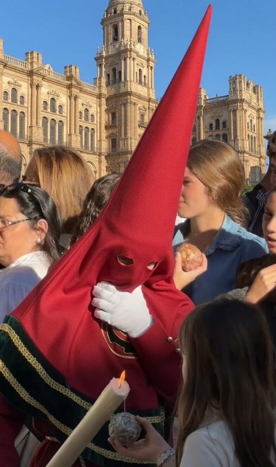 A person dressed in a traditional red Spanish 'capirote' pointed hood and white gloves at an outdoor gathering, with a historic stone building in the background and a lit candle held by someone nearby.