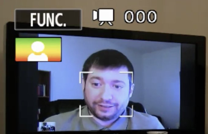 The image shows a man's face displayed on a television screen with a computer monitor behind him, both of which are black. The man appears to be smiling at the camera while looking into it. In front of the man, there is a small white box that says "Func." and has an arrow pointing towards it. A person can also be seen in the image, but they are not the main focus.