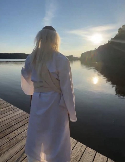 A person with long blonde hair, wearing a white robe, stands on a wooden dock looking out at a sunlit lake.