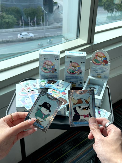 Two hands holding Pokemon trading cards, with several boxes of 'Tokocrypto' Pokemon-themed merchandise visible in the background on a table near a window.