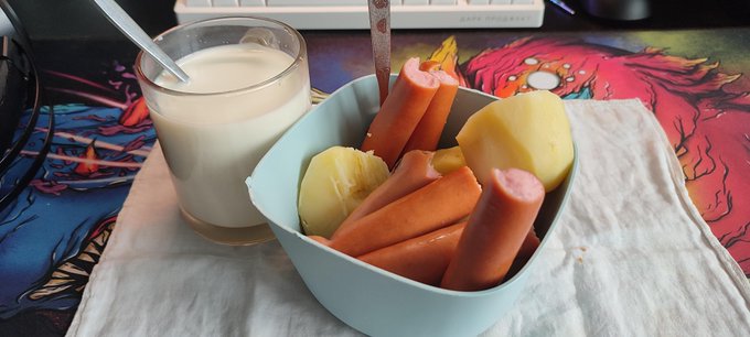 A light blue bowl holds sausages and boiled potatoes next to a glass of a light-colored beverage, sitting on a Spider-Man themed mat.