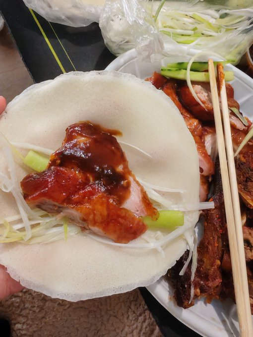 A hand holds an open Chinese pancake wrap filled with sliced green onions, cucumber, and glazed roast duck drizzled with dark brown sauce, with chopsticks resting on a foam container of additional duck meat in the background.