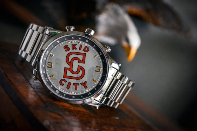 A close-up photograph of a silver chronograph wristwatch with a face displaying a red 'S' logo and the text 'SKID CITY', resting near a blurred eagle figurine.