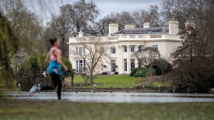 A woman in a blue sports bra and black leggings jogs past a pond in the foreground, with a large white mansion featuring columns visible across the water.