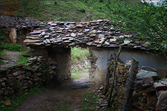 A rustic stone building with a slate roof and archway entrance sits in a green, hilly landscape.