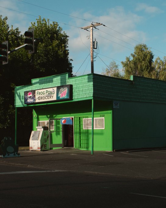 A green-painted storefront named 'Frog Pond Grocery' with neon signs, featuring Pepsi and Mountain Dew vending machines outside, located under a partly cloudy sky with trees in the background.